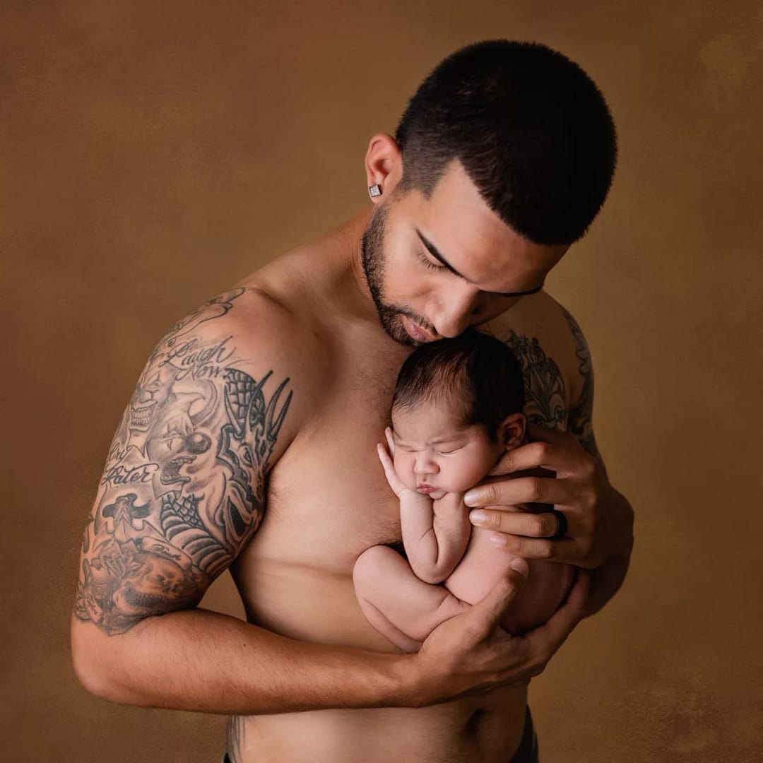 Dad posing with newborn baby against a brown backdrop. Dad is shirtless with tattooes on his arms. Baby is curled up on the chest. A sweet image with light contrast taken by San Diego Newborn Photographer in a local photography studio
