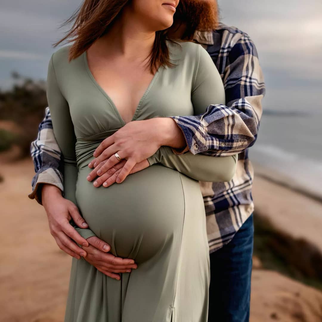 Portrait of expecting mom and dad, close up of the belly with their arms wrapped around each other. Moody feel with sunset in the background. Taken by the beach in San Diego by local maternity photographer