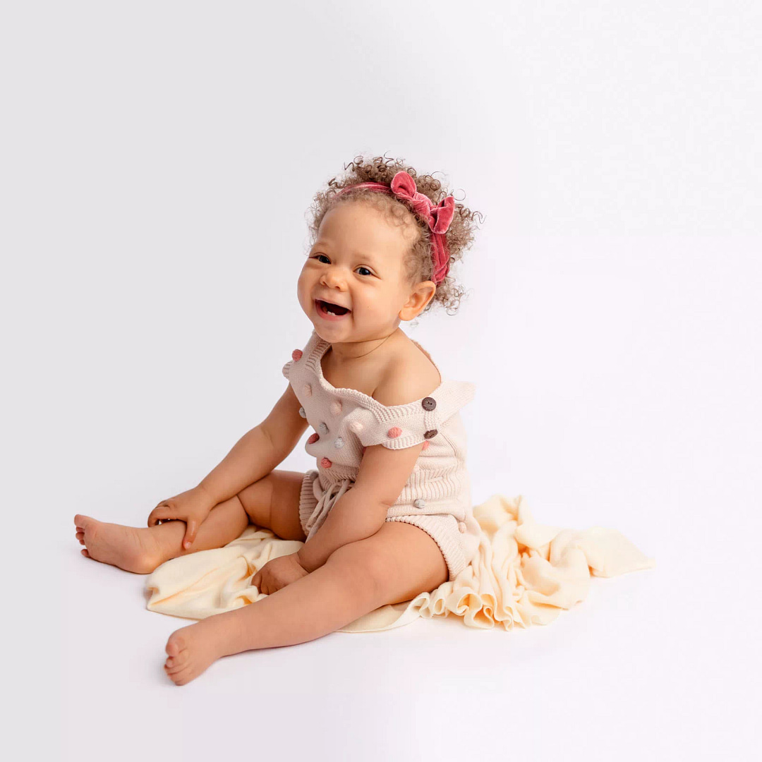 Laughing one year old girl sitting on a white floor wearing a boho outfit during her one year milestone pictures at local San Diego Portrait Studio