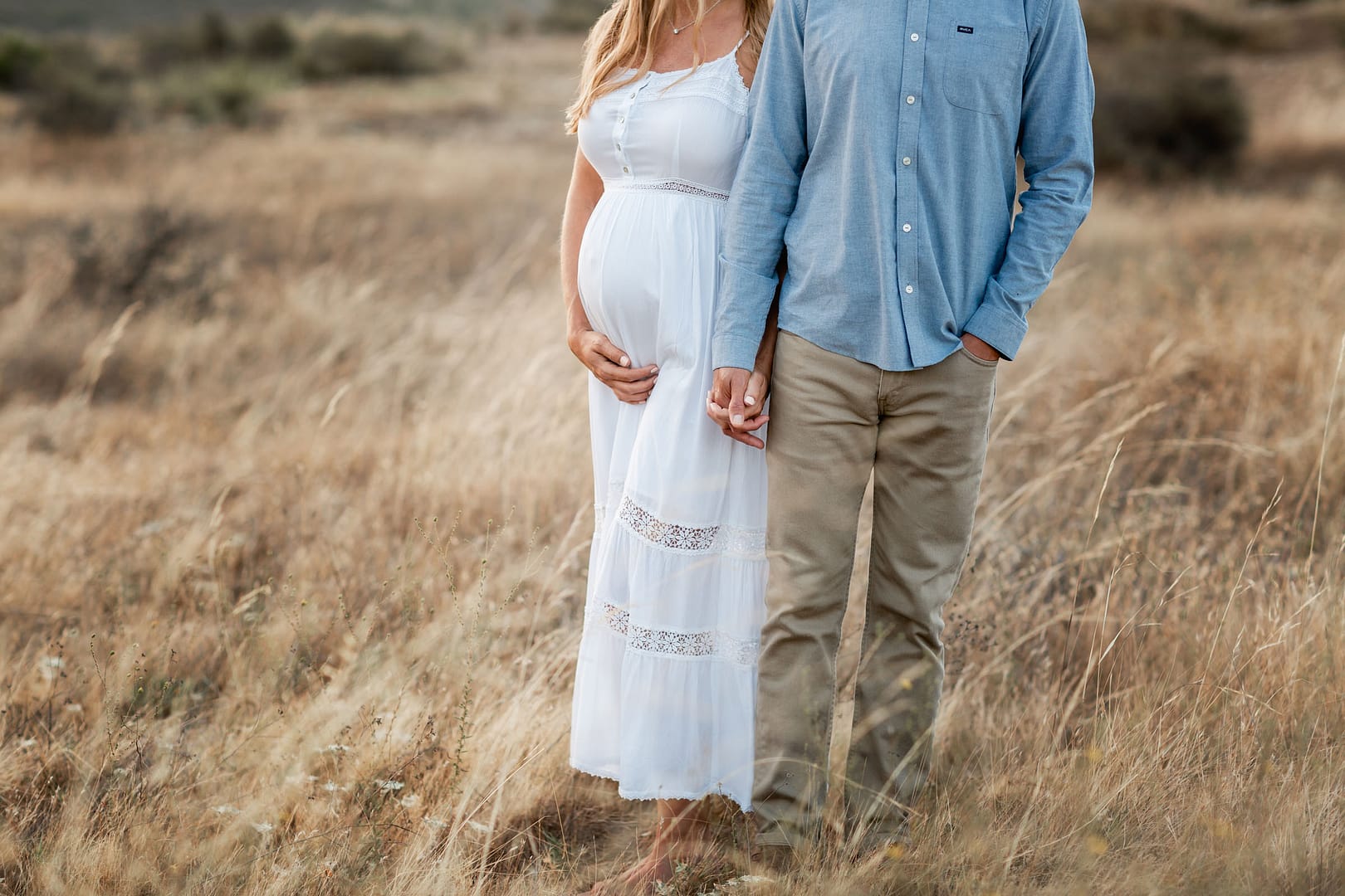 San Diego Maternity Photography of couple standing in an open field of grass, with the focus being on the belly and the couple holding hands.