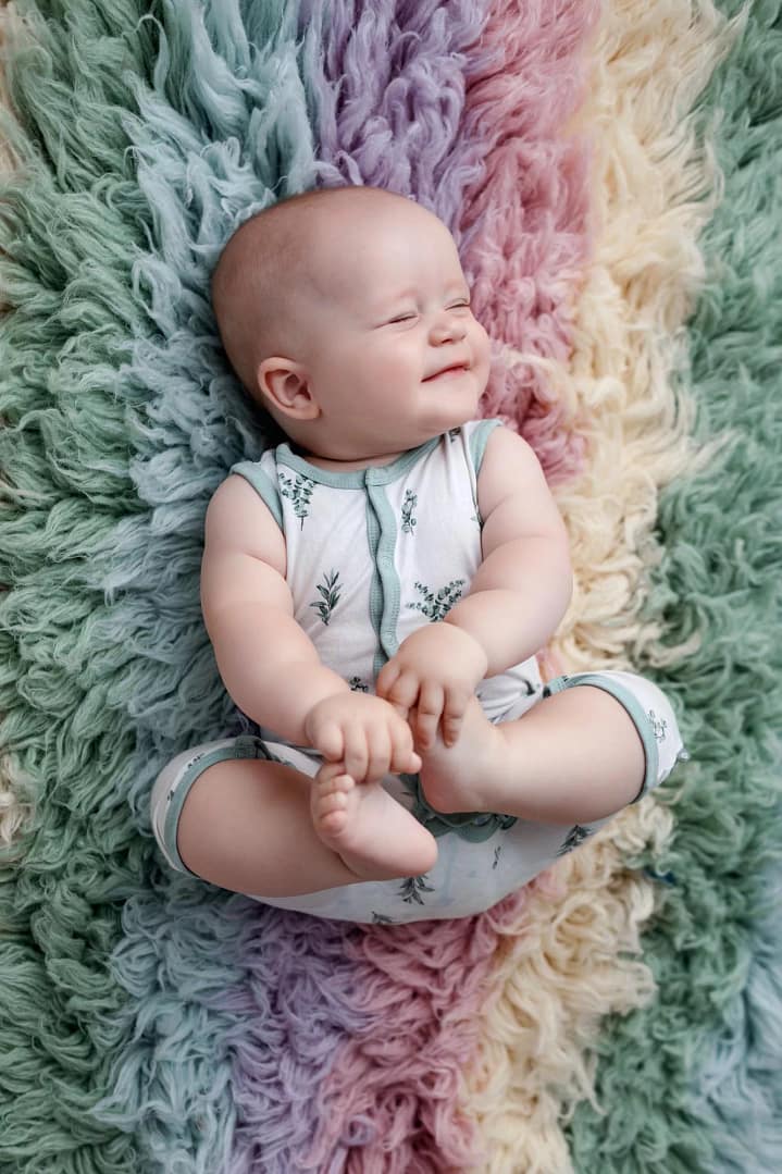 Baby 6 Month Milestone Portraits laying on a rainbow flokati rug holding on to feet and smiling with eyes closed captured by Baby Photographer in San Diego