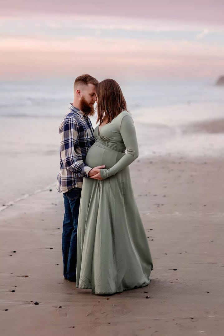 Gorgeous maternity portraits in San Diego during sunset with a pink sky, on the beach featuring mom and dad facing each other.