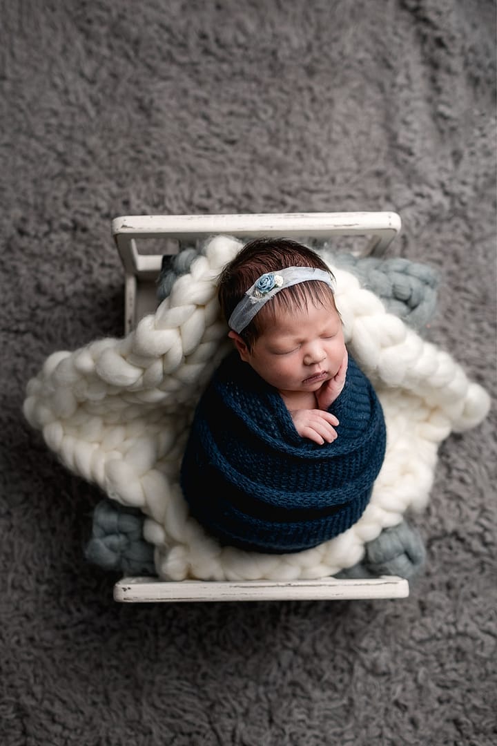 Newborn girl wrapped in blue wrap with headband laying on blankets on a newborn bed