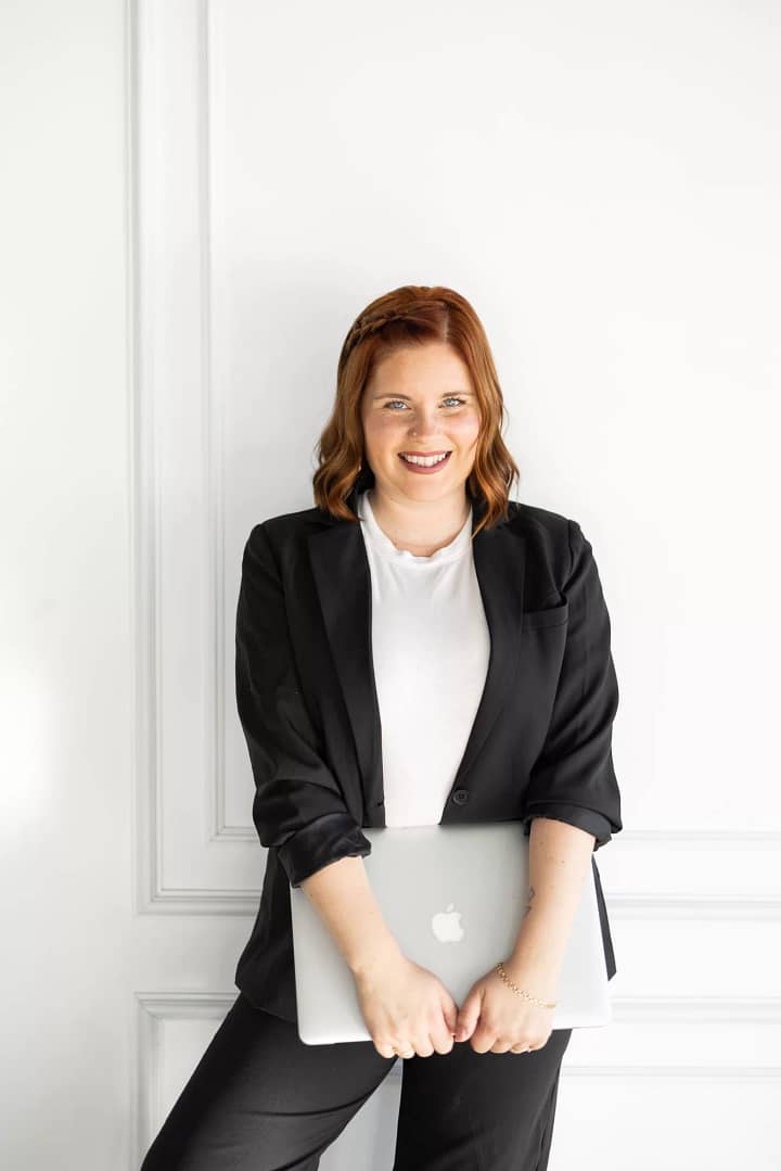 Headshot of San Diego Maternity, Newborn and Family Photographer standing against a white wall, holding her laptop wearing a white shirt and ablack blazer.