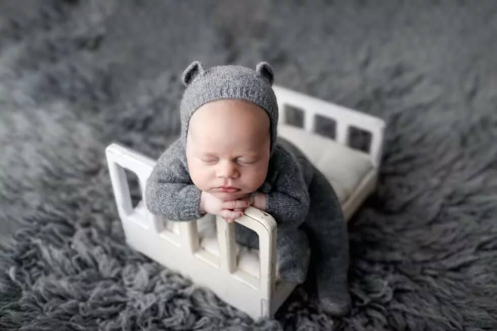 Newborn Baby Boy in a gray teddy bear suite posed on a white doll bed during his Newborn Pictures at a San Diego Professional Photography Studio