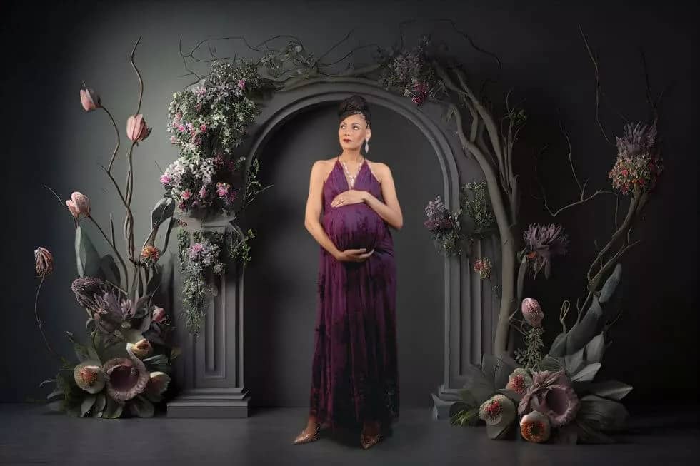 Studio Maternity Picture taken by San Diego Maternity Photographer depicting a mom in a purplish lace dress on a dark gray background with flowers and an arch