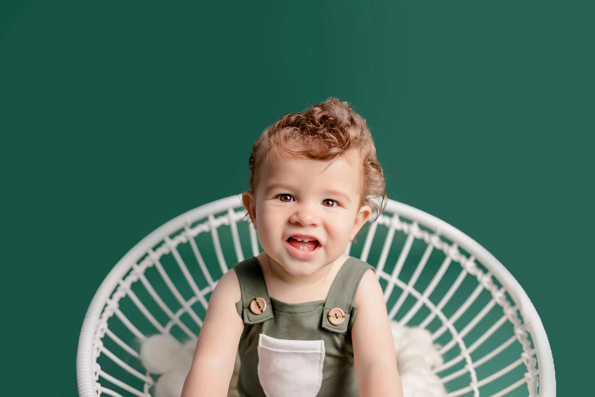 Close up of baby boy sitting in a white chair on a green backdrop wearing a green sitter outfit during milestone photos by San Diego Family Photographer Studio Freyja